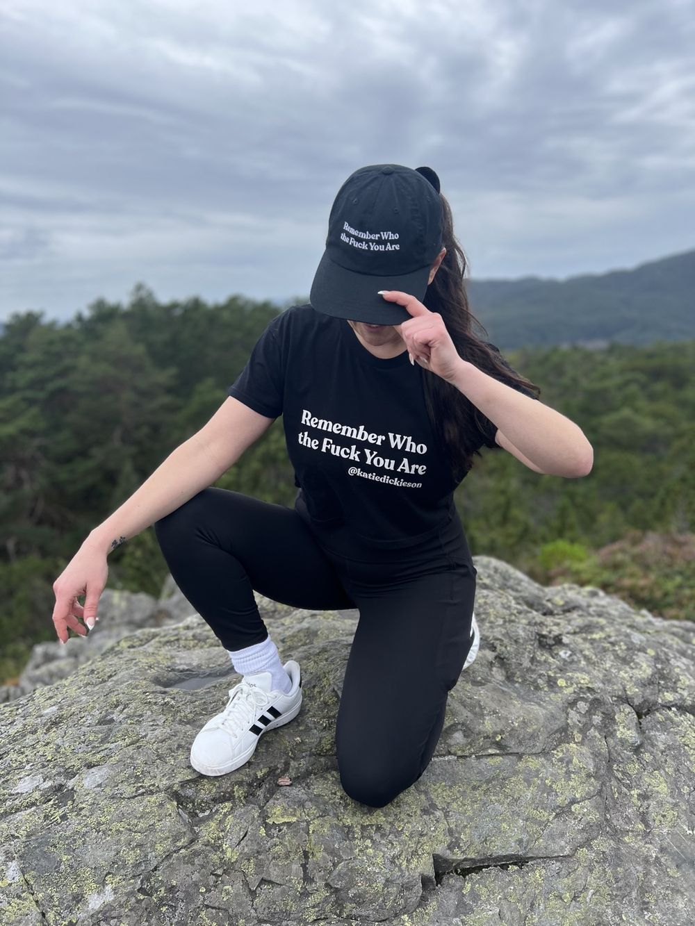 Katie Dickieson outdoors on a rock ledge, wearing a Remember Who the F*ck You Are shirt and cap.