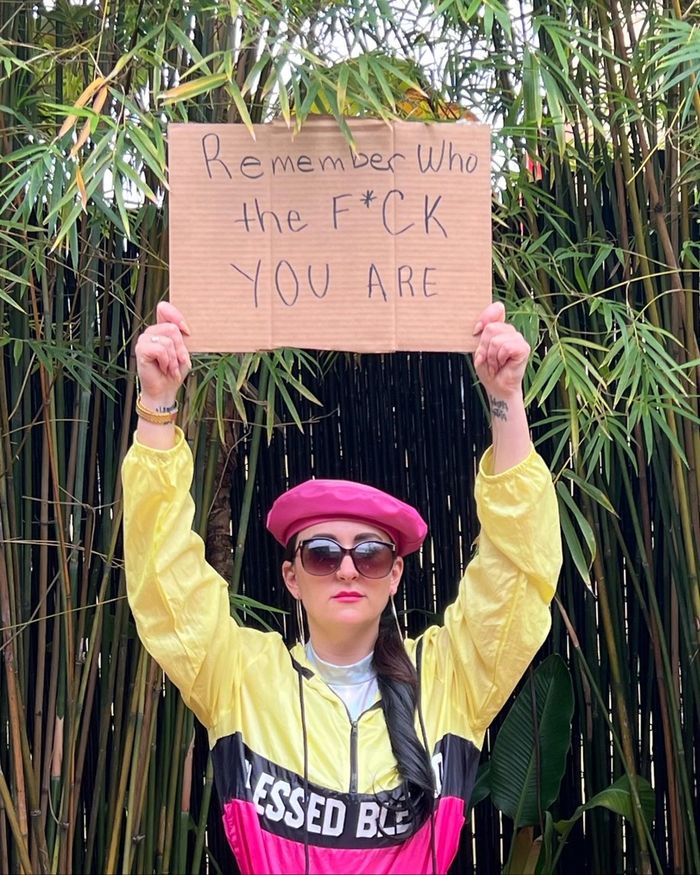 Katie Dickieson in a yellow jacket and pink beret, holding a hand-lettered cardboard sign reading Remember Who the F*CK You Are.